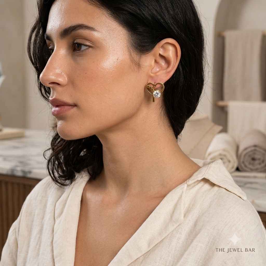 A woman in profile wearing a gold heart-shaped stud earring with a clear central gemstone, set against a softly blurred spa-like background. The text 'THE JEWEL BAR' is visible in the bottom right corner.