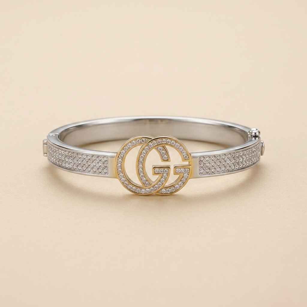 A silver-toned Gucci bangle bracelet featuring a central gold-toned interlocking GG logo encrusted with small clear stones, set against a light beige background.
