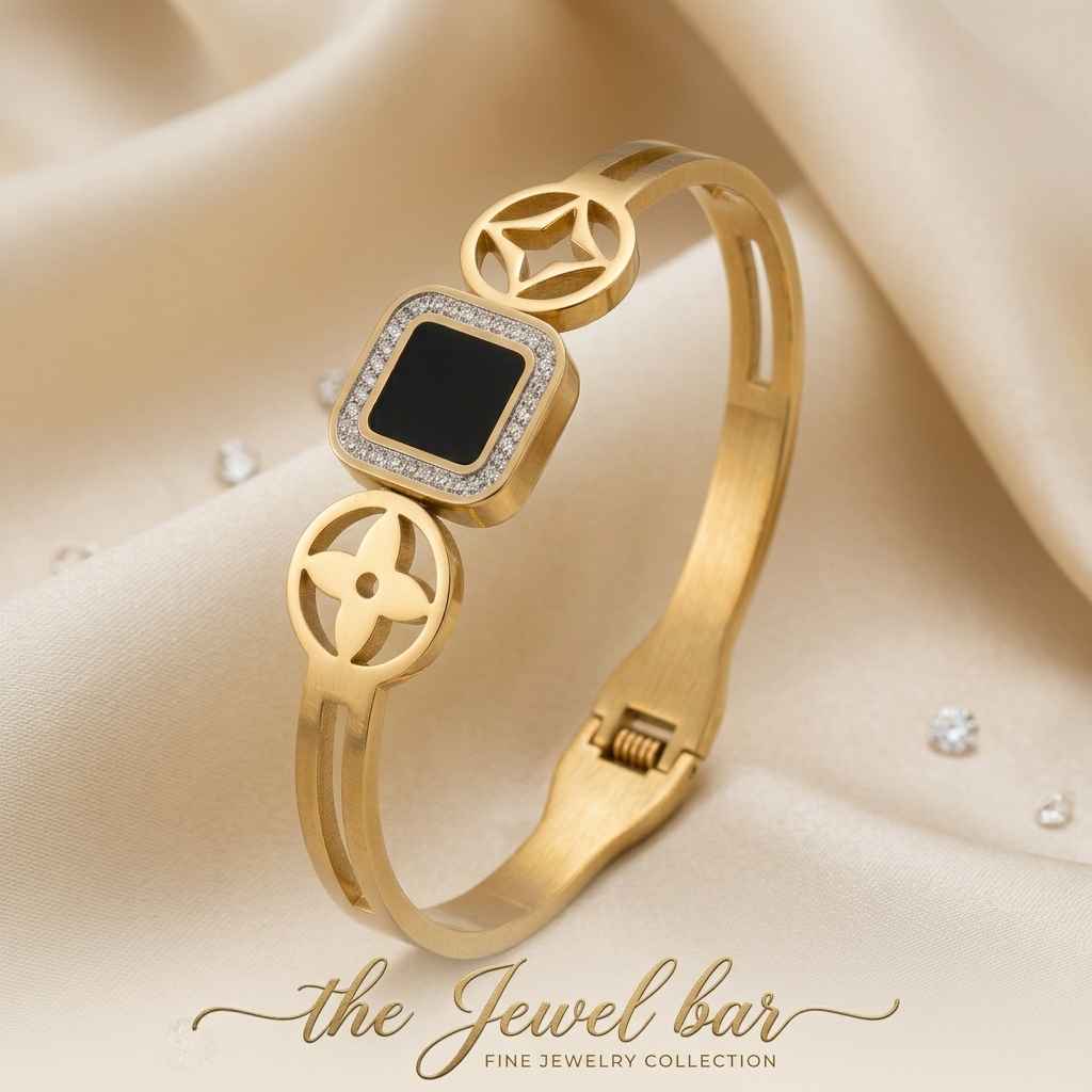 A gold-plated bangle bracelet with a central square black inlay bordered by small clear stones, displayed on draped cream satin fabric with a 'the Jewel bar' watermark at the bottom.