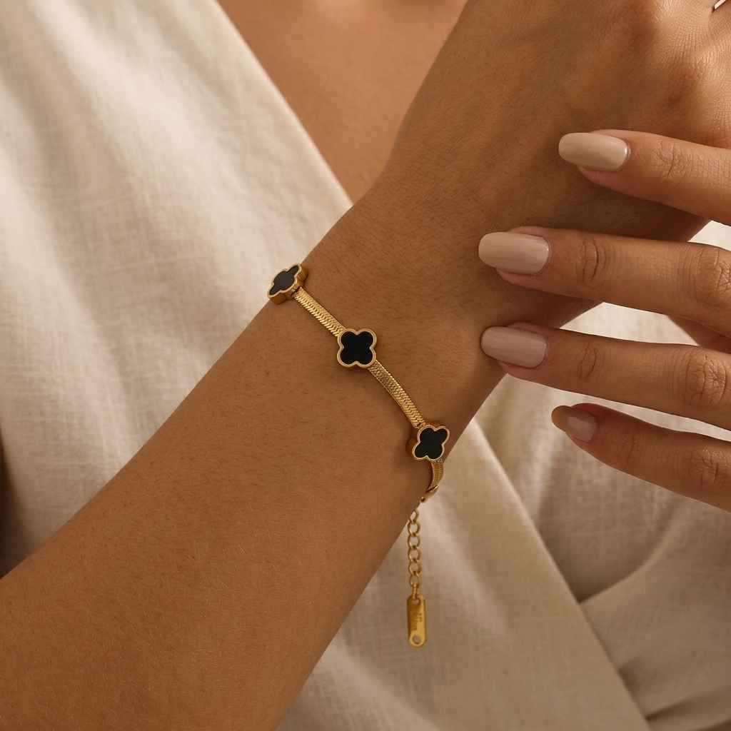 A gold-tone snake chain bracelet with three black enameled four-leaf clover motifs, shown worn on a woman's wrist with her fingers resting nearby.