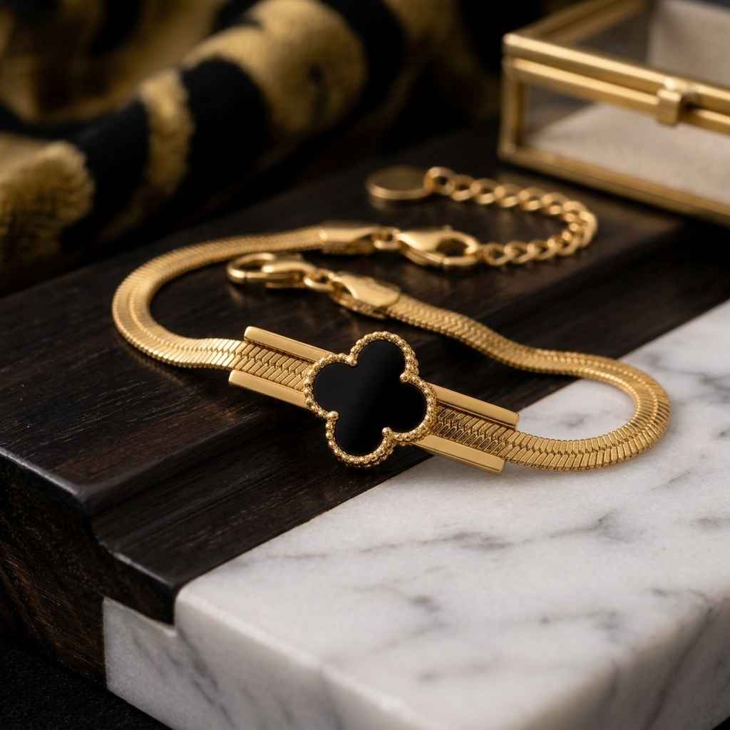 A gold-plated snake chain bracelet with a black enamel four-leaf clover motif and a polished gold bar detail, displayed on a dark wood block and white marble surface.
