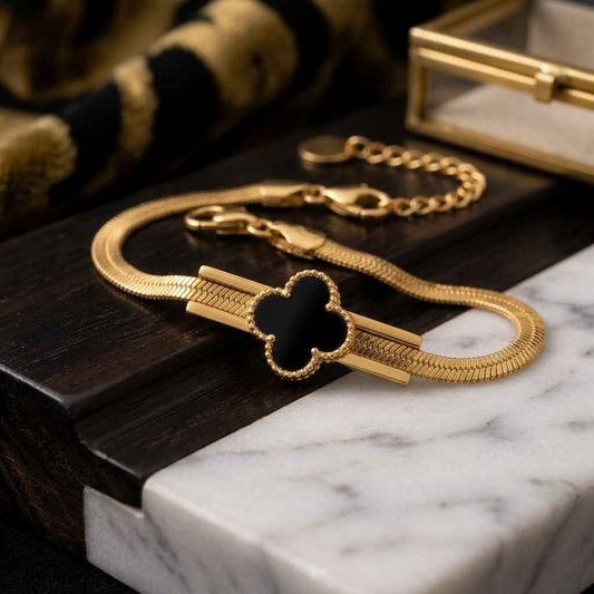 A gold-plated snake chain bracelet with a black enamel four-leaf clover motif and a polished gold bar detail, displayed on a dark wood block and white marble surface.
