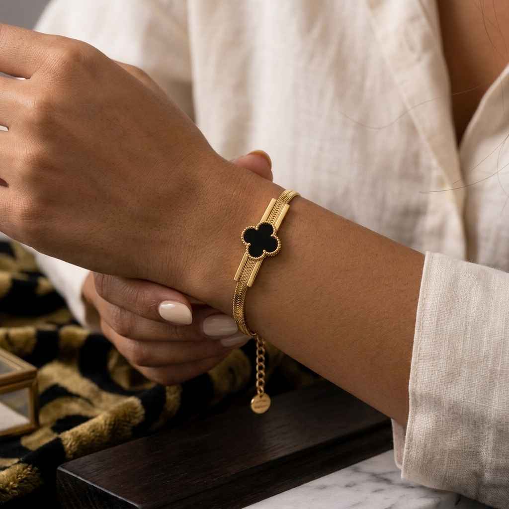 A close-up of a person's wrist wearing a gold-tone chain bracelet with a black four-leaf clover charm, paired with a white linen shirt.