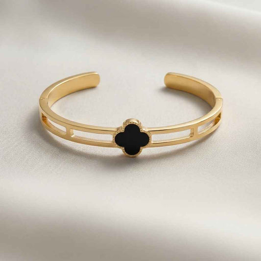 A gold-tone open cuff bracelet featuring a central black clover-shaped inlay, displayed on a soft, light-colored fabric background.