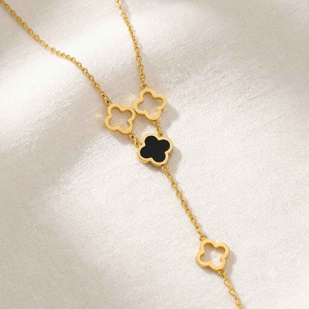 A delicate gold chain necklace featuring four clover-shaped charms, with one charm filled in black, displayed on a soft white fabric background.