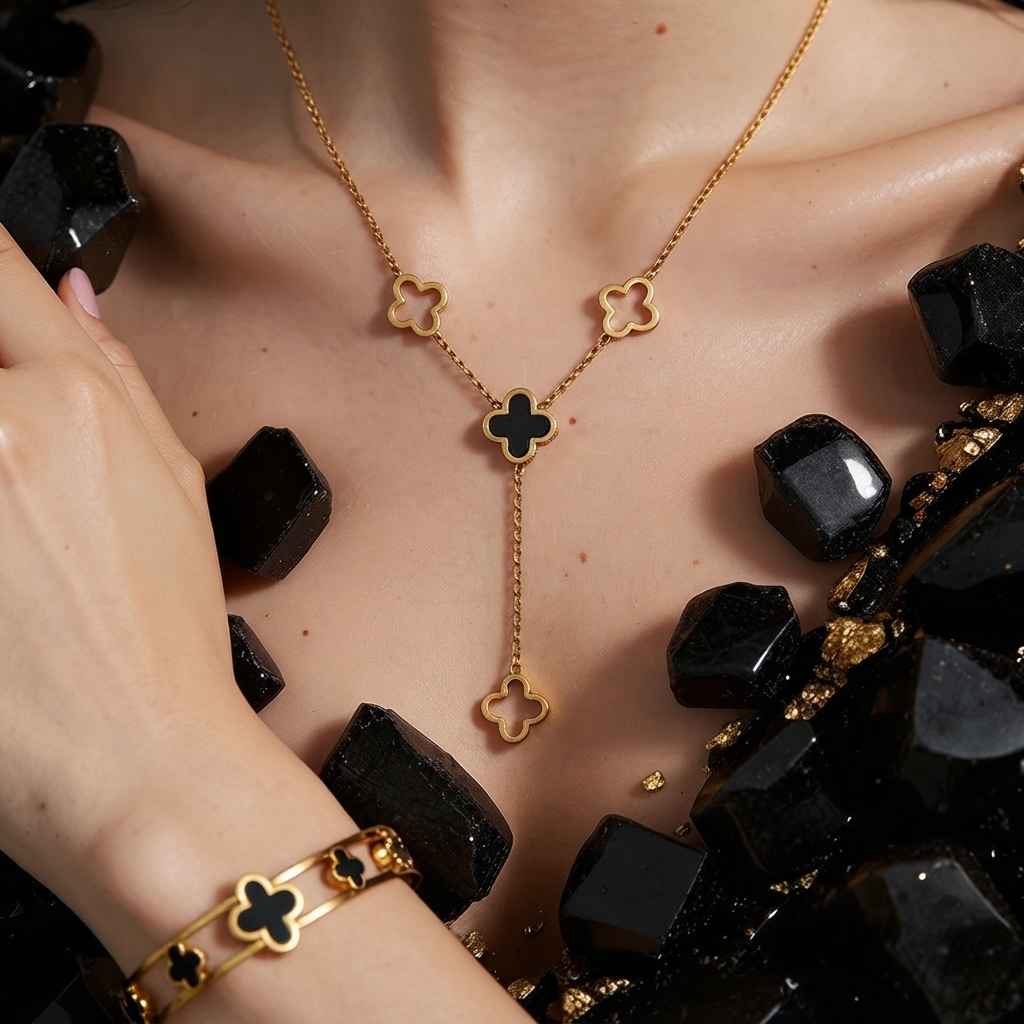 Gold-toned jewelry set featuring a Y-necklace and matching bracelet with clover motifs, worn on a model's neck and wrist, surrounded by black crystal-like stones.