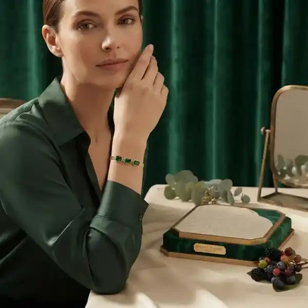 A woman in a dark green satin shirt rests her head on her hand, wearing a gold bracelet with green rectangular stones. In front of her on a cream tablecloth is a green velvet jewelry box with a gold plaque, a small mirror, and greenery, set against a dark green velvet curtain.