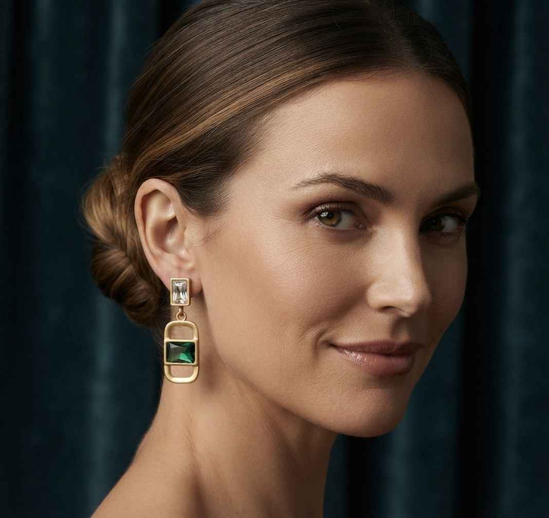 A close-up portrait of a woman wearing a gold drop earring with a rectangular green stone, set against a dark blue velvet curtain background.