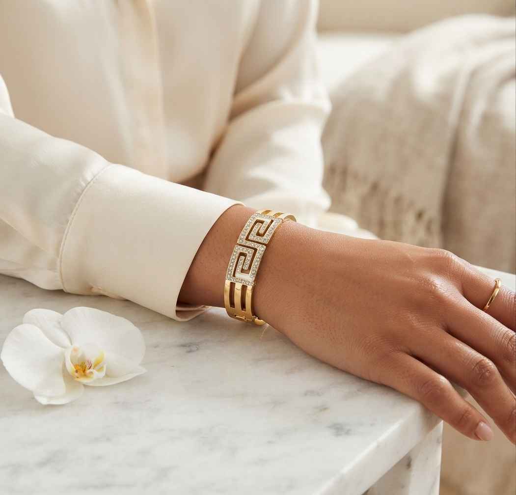A gold-tone cuff bracelet with a Greek key pattern and pavé-set clear stones, worn on a woman's wrist resting on a white marble surface.