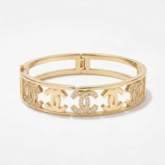 A gold-tone metal bangle bracelet featuring four interlocking Chanel CC logos, some embellished with small clear crystals, set against a plain white background.