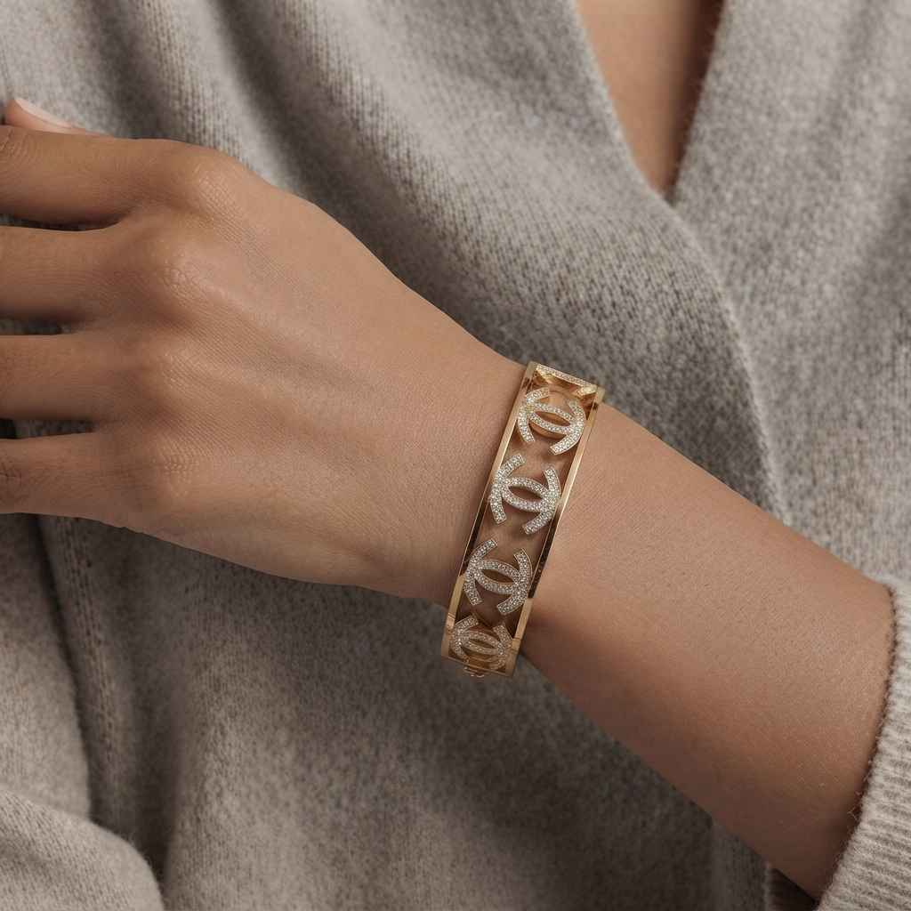 A gold-toned bangle bracelet with a repeating interlocking double-C logo pattern, worn on a person's wrist over a taupe knit sweater.