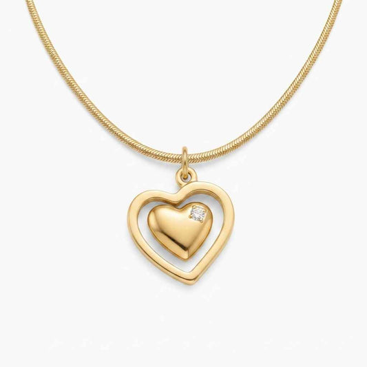 A gold heart pendant necklace featuring a polished inner heart with a small clear gemstone, set within an open heart-shaped frame on a delicate gold chain, shown against a light gray background.