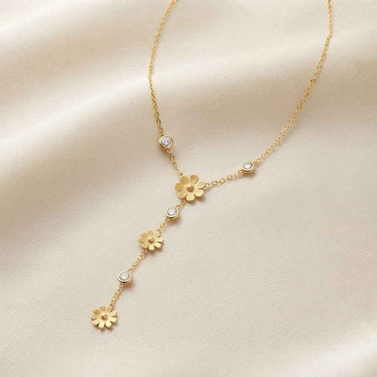 A delicate gold Y-shaped necklace with small flower charms and clear rhinestone accents, displayed on a soft beige satin fabric background.