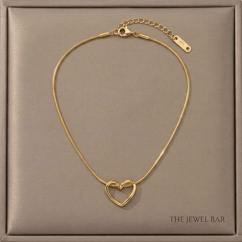 A gold heart-shaped chain necklace with a small interlocking heart pendant, displayed on a textured taupe jewelry box. The brand name 'THE JEWEL BAR' is printed in the bottom right corner of the box.