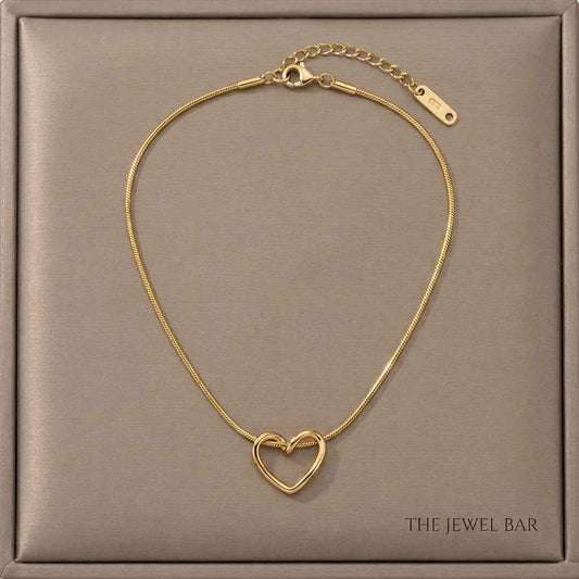 A gold heart-shaped chain necklace with a small interlocking heart pendant, displayed on a textured taupe jewelry box. The brand name 'THE JEWEL BAR' is printed in the bottom right corner of the box.