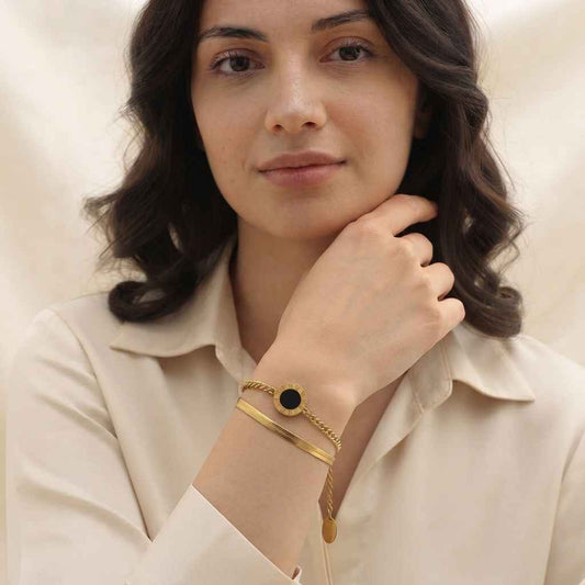 A woman wearing a delicate gold bracelet with a round black inlay, styled with a thin gold chain bracelet, against a soft beige background.