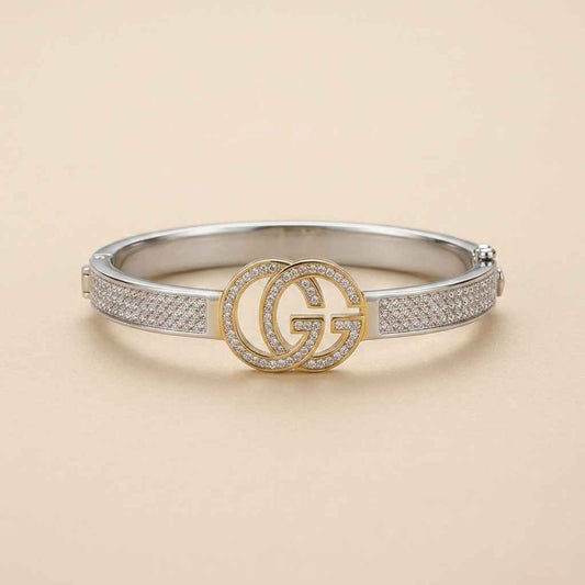 A silver-toned Gucci bangle bracelet featuring a central gold-toned interlocking GG logo encrusted with small clear stones, set against a light beige background.