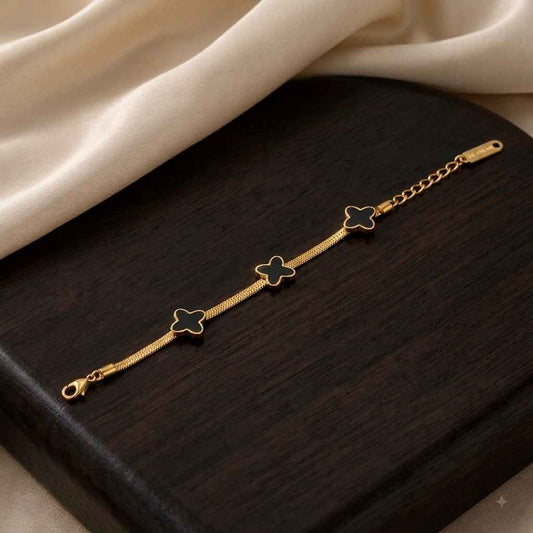 A gold chain bracelet with three clover motifs, each filled with black enamel, displayed on a dark wooden block with cream-colored fabric in the background.