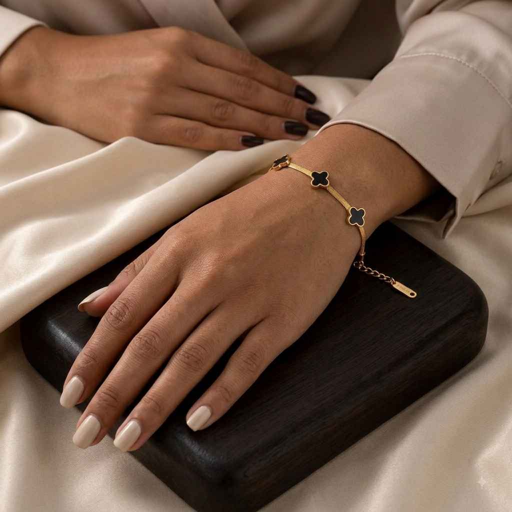 A gold bracelet with three clover motifs, two featuring black enamel centers, worn on a woman's wrist. The woman's hands with manicured nails rest on a dark wooden box over a cream satin fabric background.