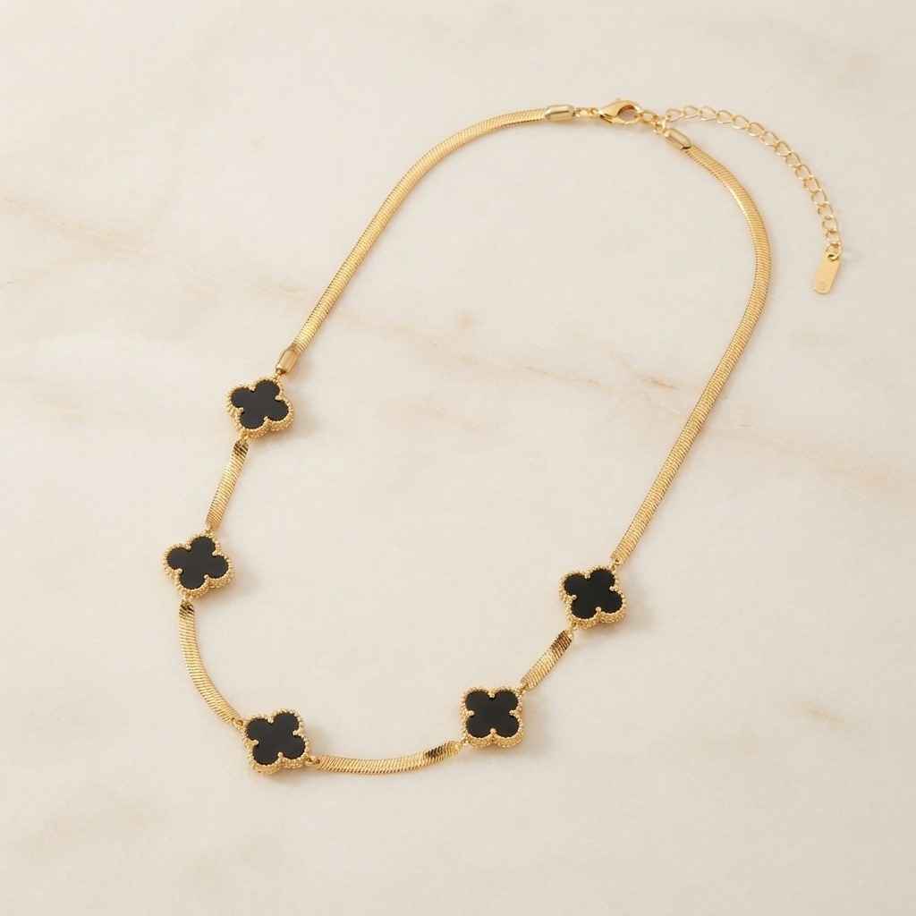 A gold chain necklace featuring five black clover-shaped charms, displayed on a light beige marble surface.