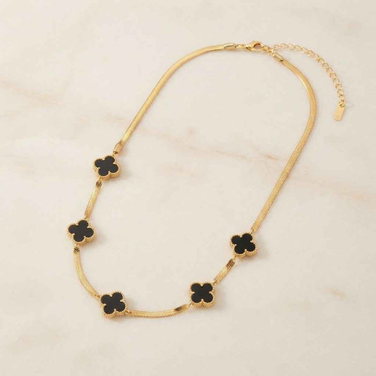 A gold chain necklace featuring five black clover-shaped charms, displayed on a light beige marble surface.