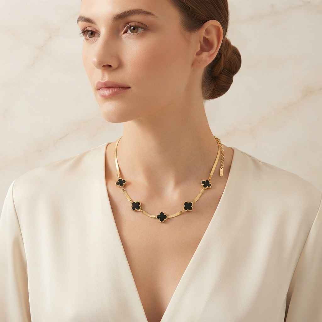 A woman wearing a gold-toned necklace with five black clover motifs, styled with a cream satin top against a light marble background.