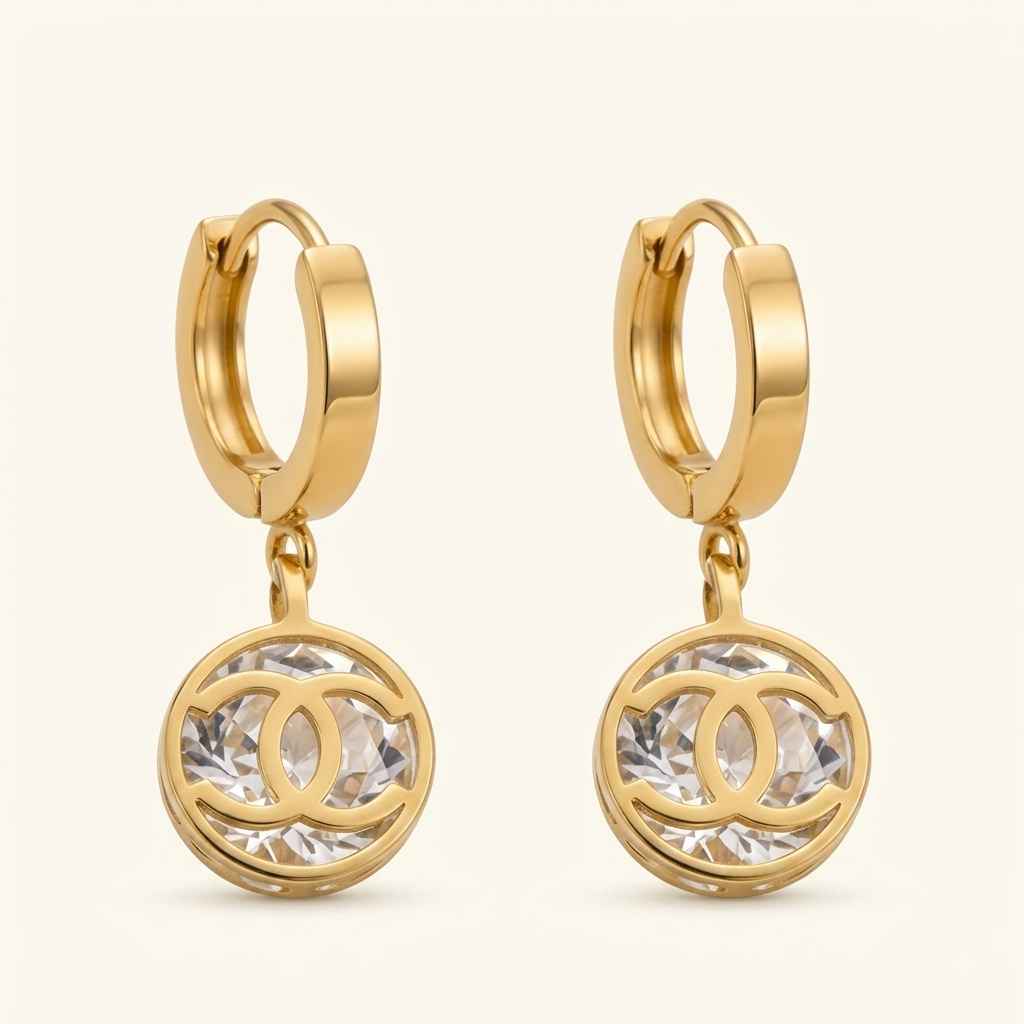 A pair of gold huggie hoop earrings with round drop charms featuring a clear faceted crystal interlocking CC logo, set against a light cream background.