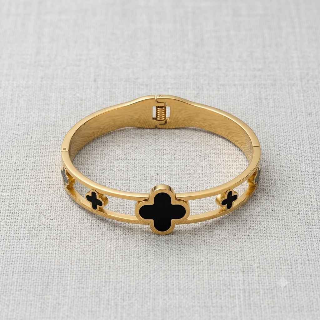 A gold-plated bangle bracelet with black enamel clover motifs, displayed on a textured light gray fabric background.