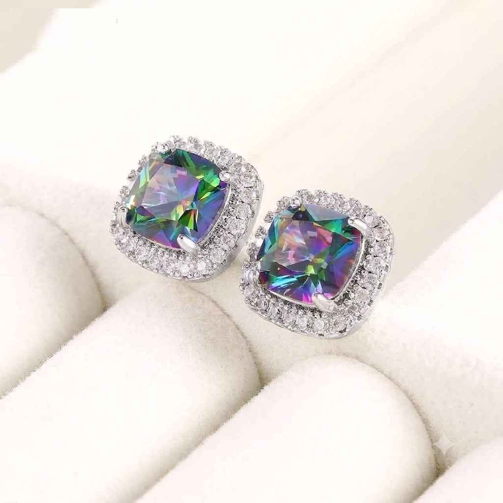 A pair of silver stud earrings with square iridescent green and purple cabochon center stones surrounded by halo clear crystals, displayed on a white velvet jewelry box.