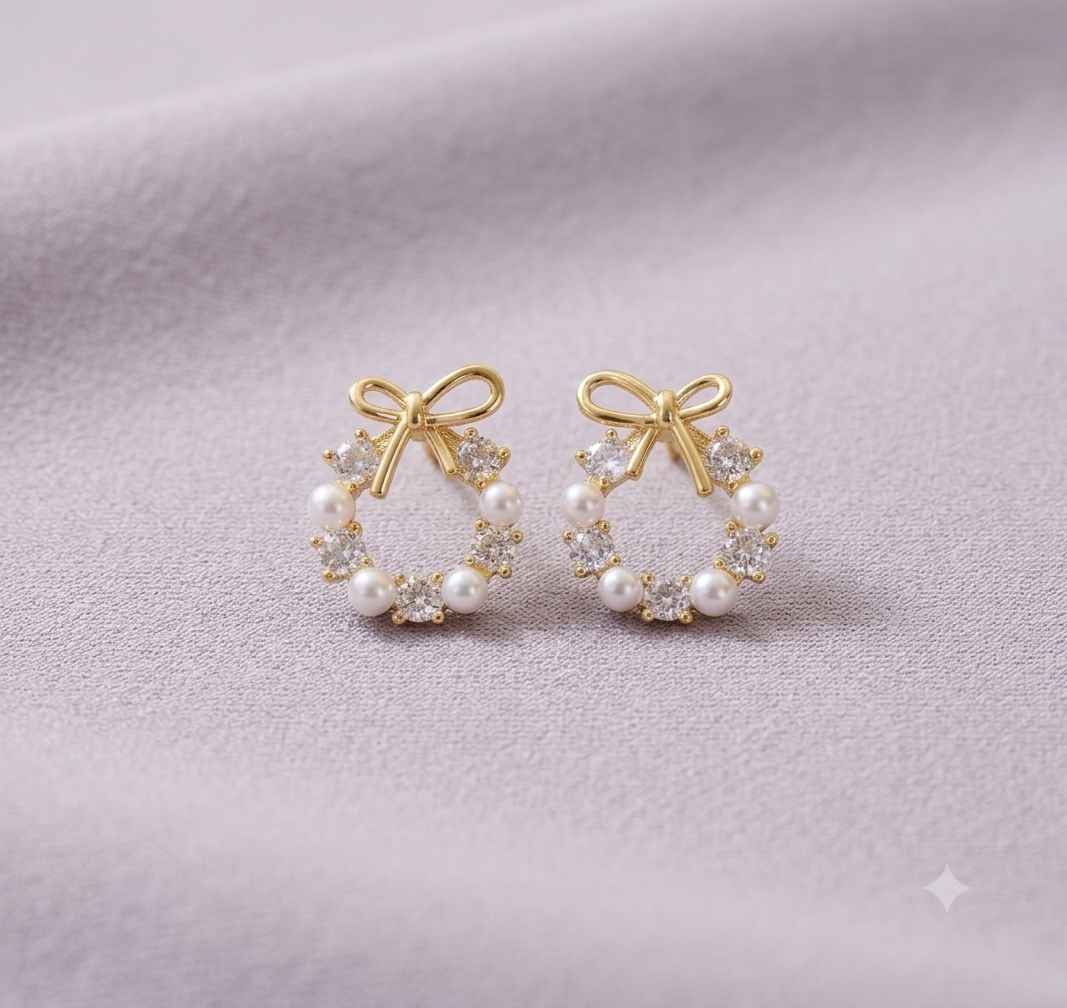 A pair of gold hoop earrings with white pearl accents and clear crystal bow details, displayed on a soft lavender fabric background.