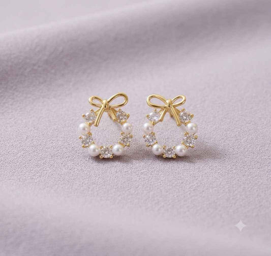A pair of gold hoop earrings with white pearl accents and clear crystal bow details, displayed on a soft lavender fabric background.