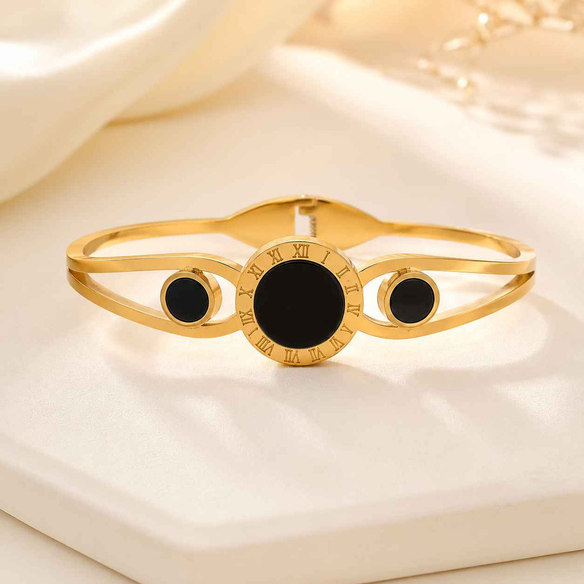 Gold-tone bangle bracelet with a central black oval stone and two smaller black oval stones, accented with Roman numerals around the center ring, displayed on a cream-colored surface.