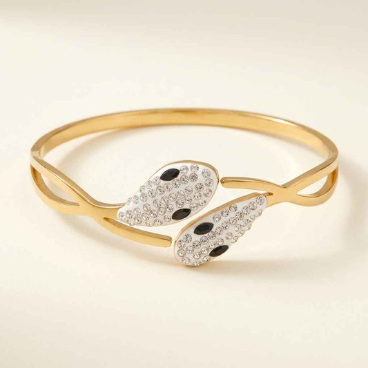 Gold-tone open cuff bangle bracelet featuring two stylized wing-like charms paved with clear rhinestones and accented with two black oval stones each, shown on a warm off-white background.