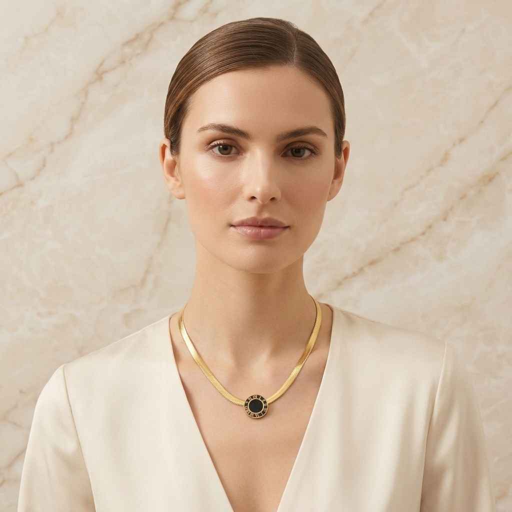 A woman with her hair pulled back wears a delicate gold chain necklace with a circular black and gold pendant, set against a light beige marble wall.