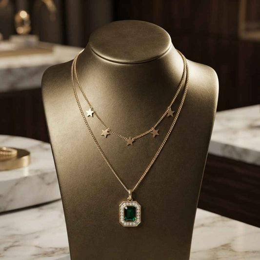A gold necklace displayed on a dark metallic jewelry bust, featuring a square emerald-cut green gemstone pendant surrounded by small clear stones, with two delicate gold star chains above it.
