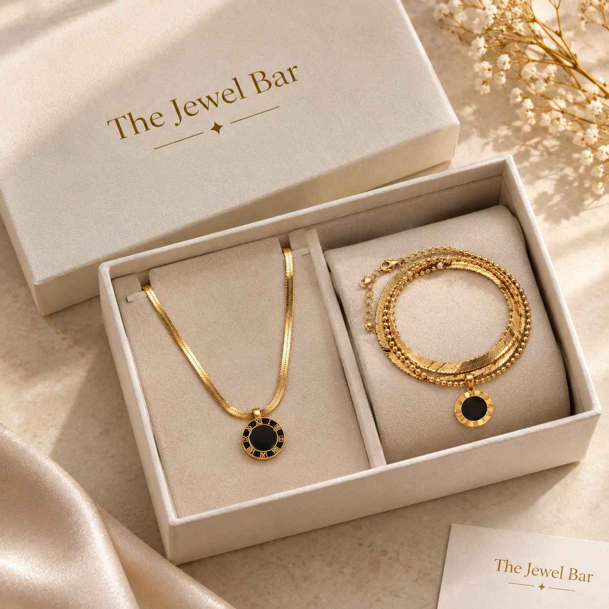 Gold jewelry set from The Jewel Bar, featuring a necklace and a multi-strand bracelet with black circular accents, presented in a cream-colored gift box with the brand name printed on the lid.