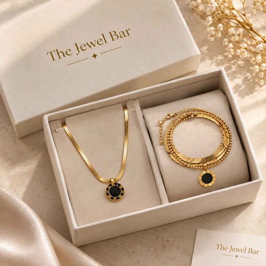 Gold jewelry set from The Jewel Bar, featuring a necklace and a multi-strand bracelet with black circular accents, presented in a cream-colored gift box with the brand name printed on the lid.