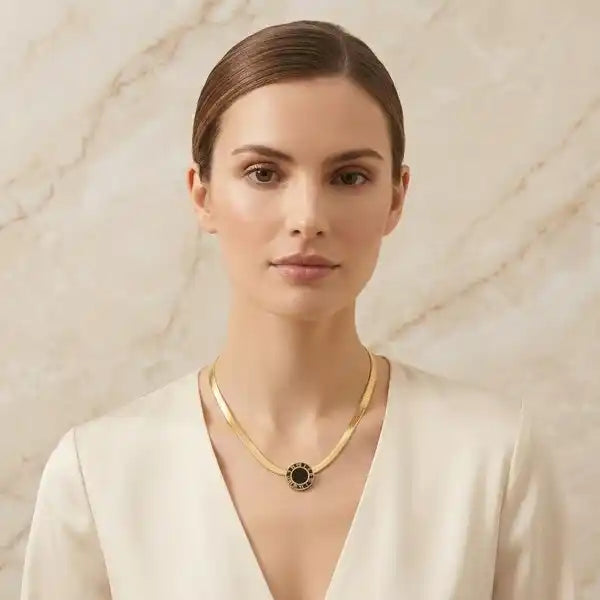 A woman with her hair pulled back wears a delicate gold chain necklace with a circular black and gold pendant, set against a light beige marble background.